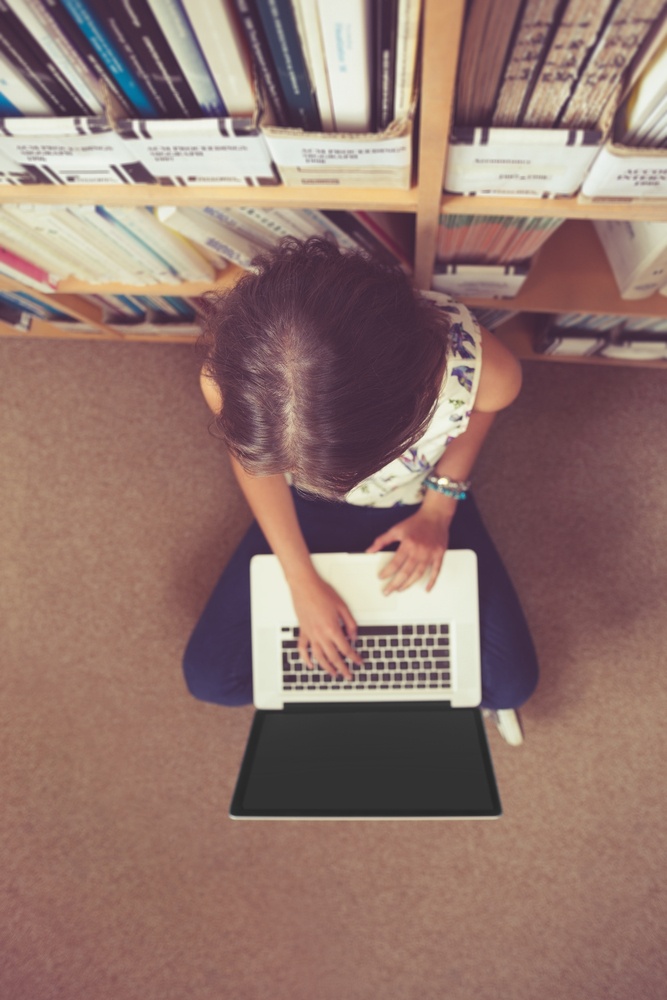 High angle view of a female student sitting against bookshelf and using laptop on the library floor
